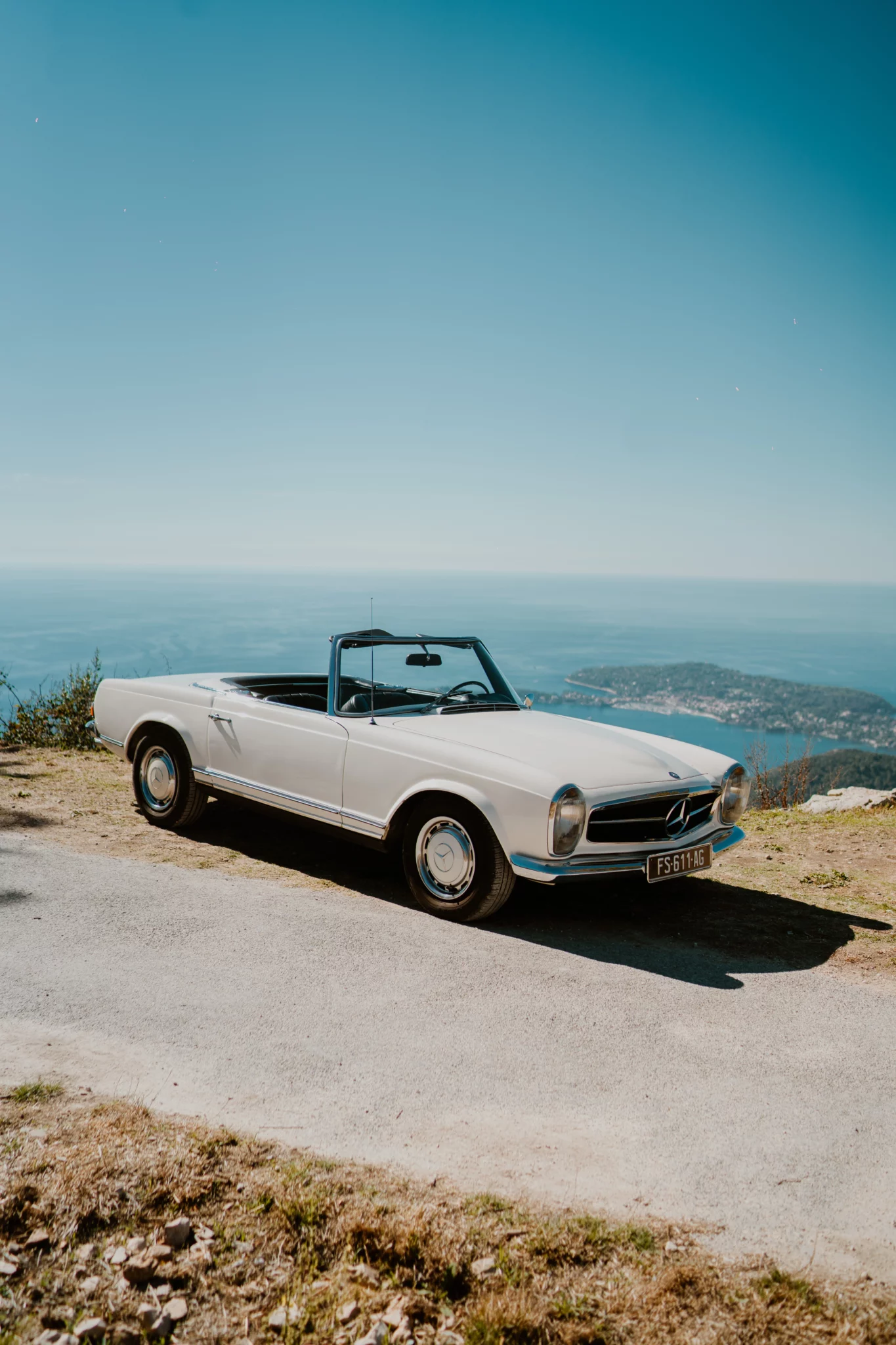 A classic white Mercedes 280SL Pagode convertible parked on a scenic coastal road overlooking the Mediterranean sea.