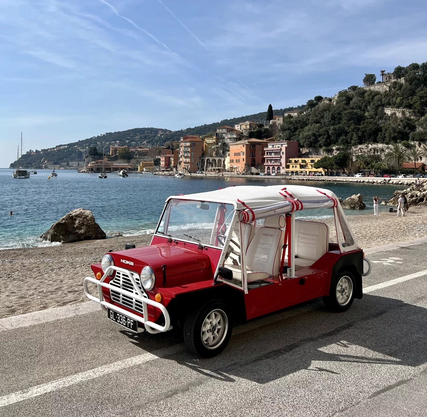 A red 1970 Mini Moke parked on a road next to a beach with the sea and a coastal town in the background.