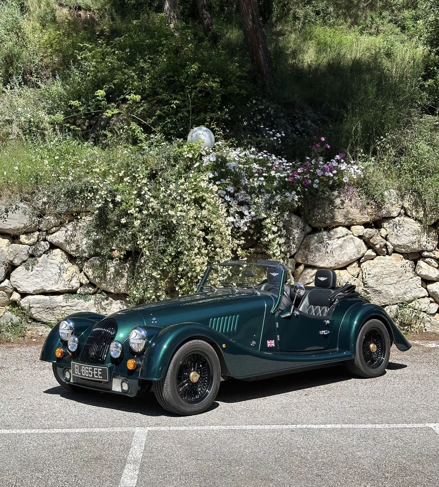 Front three-quarter view of the Morgan Plus Four in a parking spot with a stone wall behind it.
