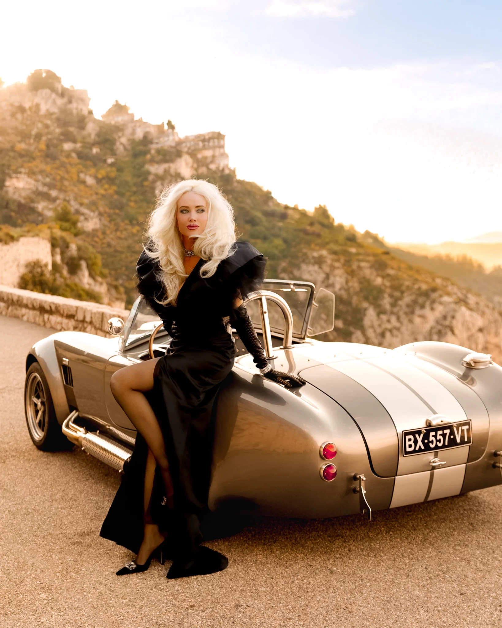 Glamorous woman with blonde hair in a black dress posing elegantly on a gray Shelby Cobra classic car on a mountain road overlooking a medieval village.