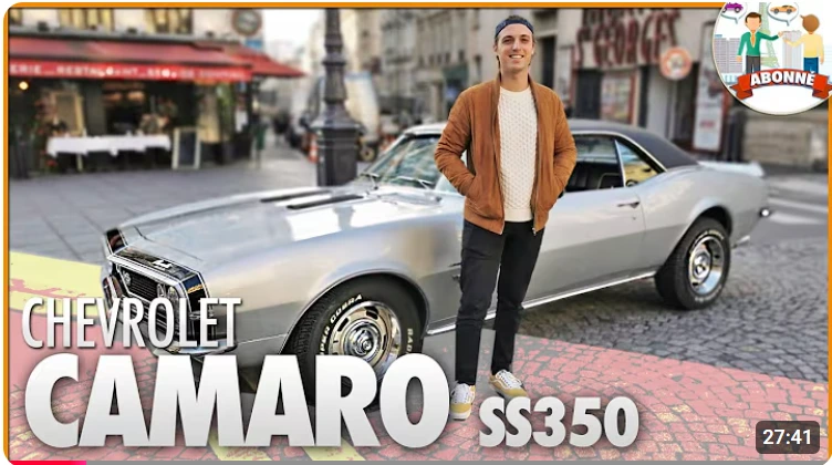Man standing in front of a silver Chevrolet Camaro SS350 classic car on a city street.