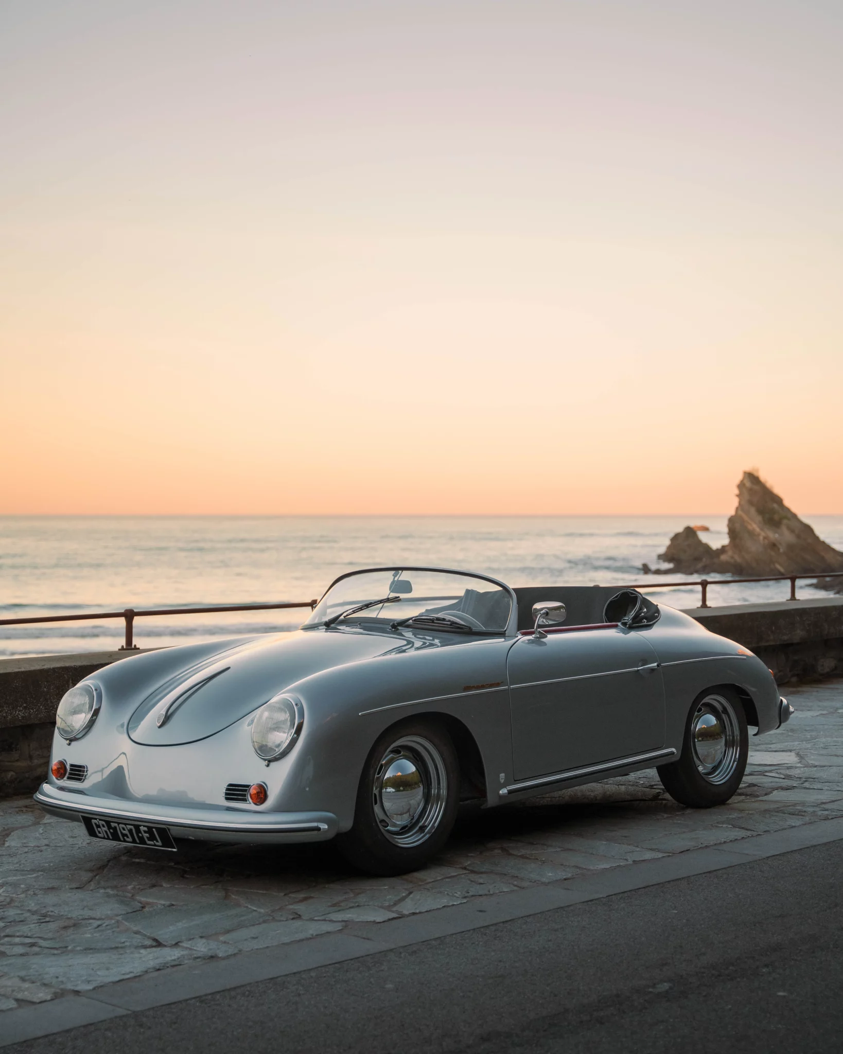 A grey 1970 Porsche 356 Speedster vintage car parked on a stone promenade with the ocean at sunset.
