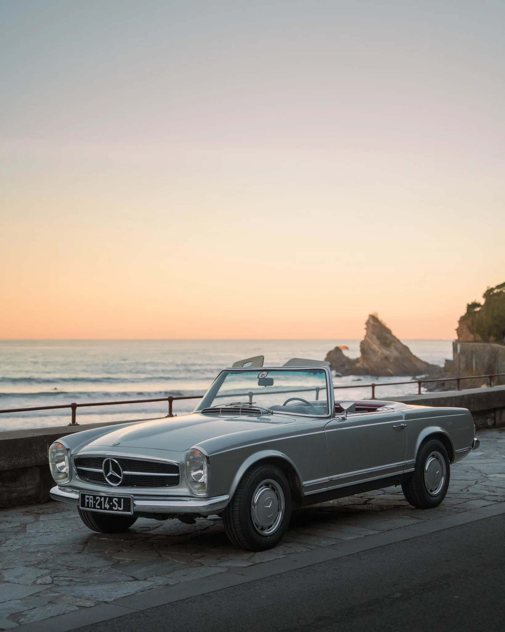 A silver 1968 Mercedes 280SL Pagoda convertible parked on a coastal promenade at sunset.