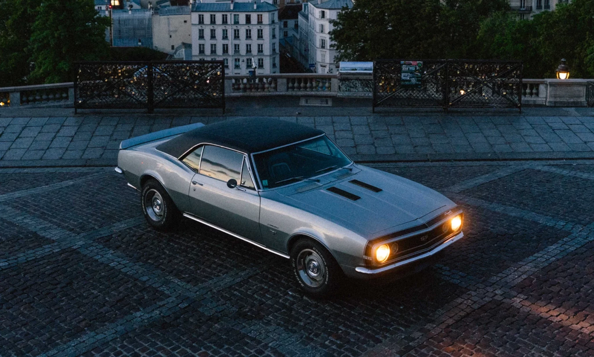 Silver classic Chevrolet Camaro parked on a cobbled street overlooking the Paris skyline at dusk.