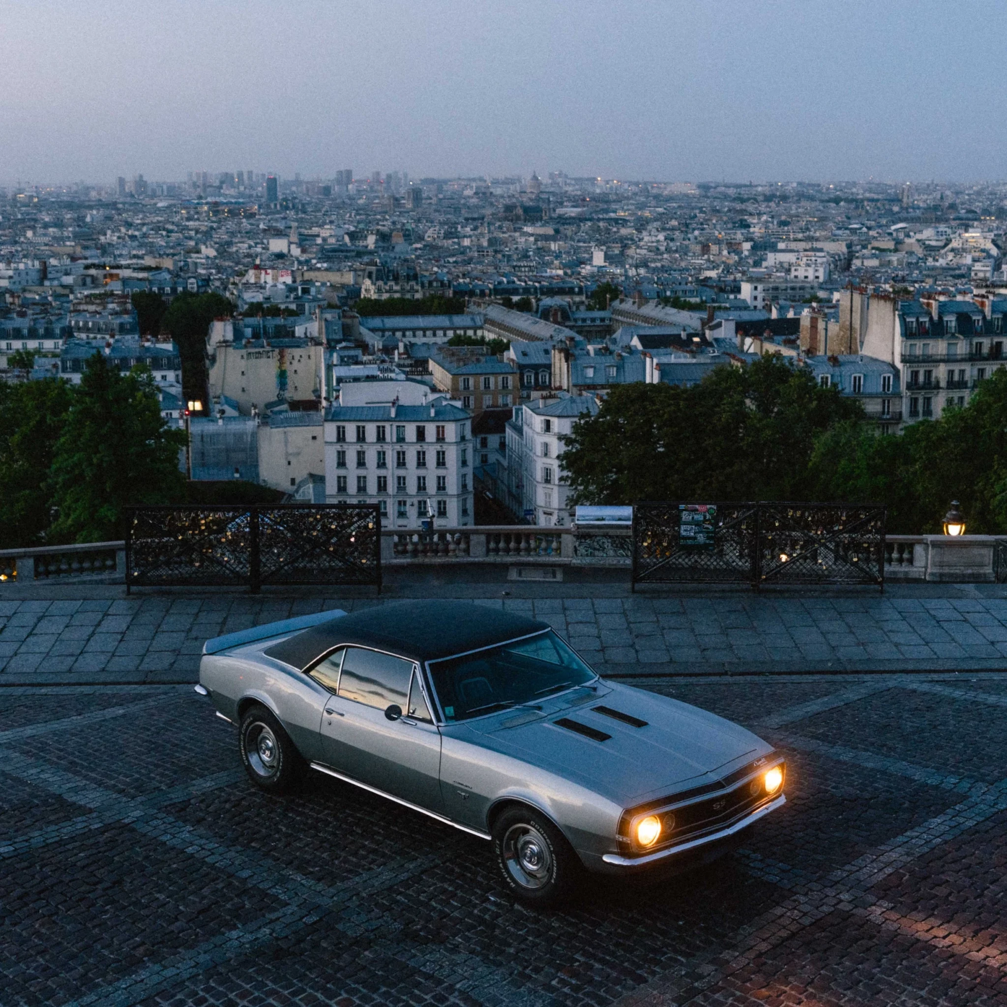Silver classic Chevrolet Camaro parked on a cobbled street overlooking the Paris skyline at dusk.
