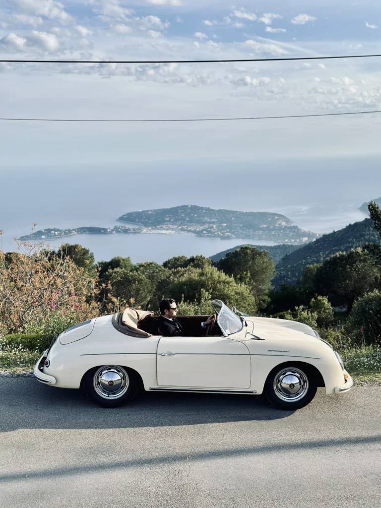Man driving a cream classic Porsche Speedster convertible on a mountain road overlooking the sea.