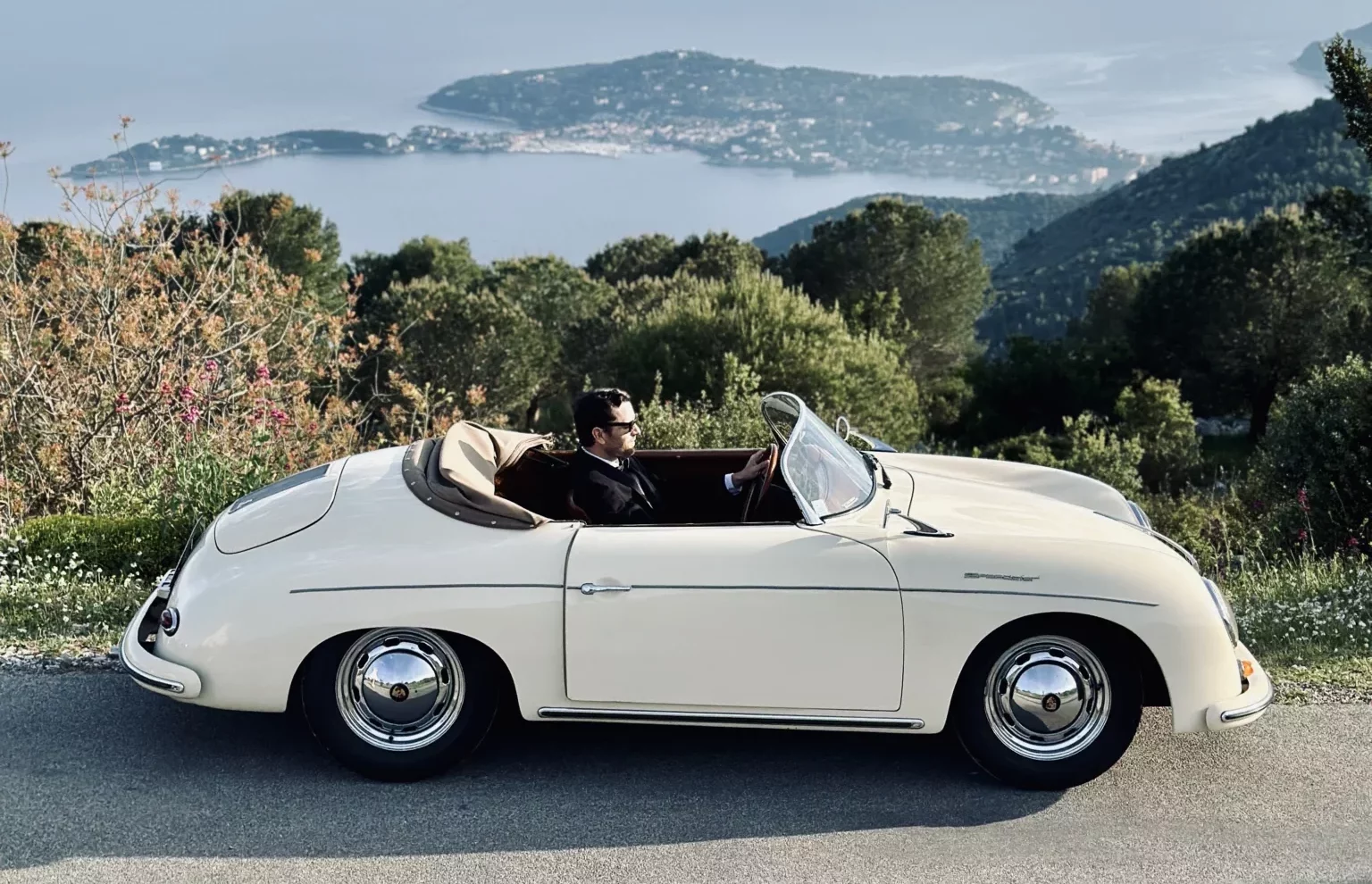 Man driving a cream classic Porsche Speedster convertible on a mountain road overlooking the sea.