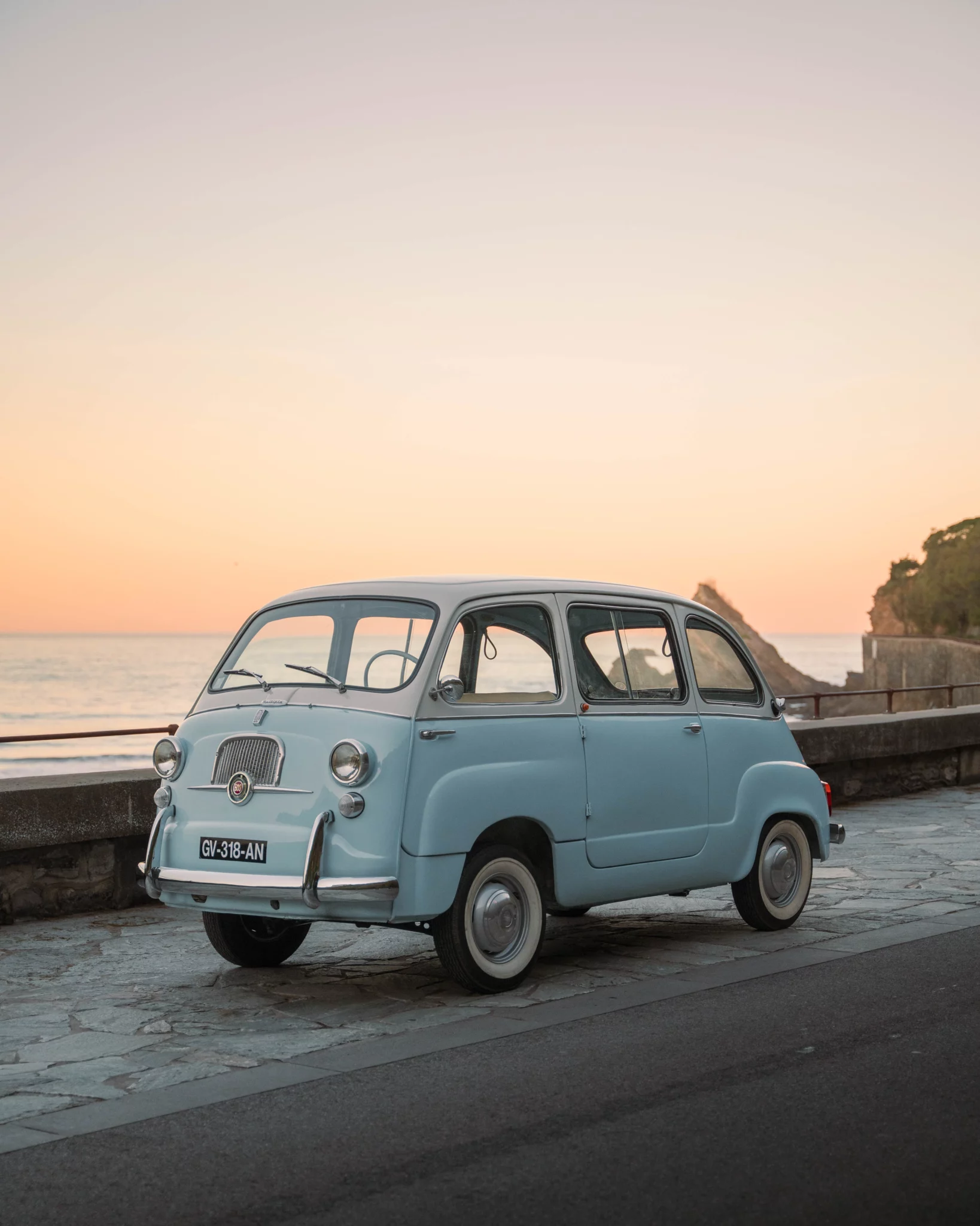 A light blue 1963 Fiat 600 Multipla vintage car parked with the ocean and a sunset in the background.