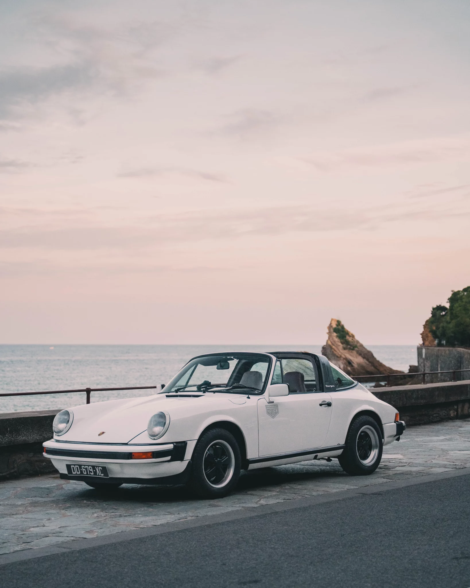 White 1977 Porsche 911 Targa vintage sports car parked on a promenade overlooking the ocean at sunset.