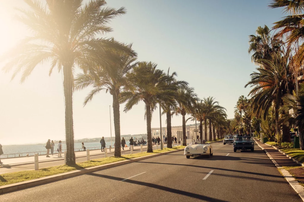 Vintage cars driving along the palm-lined Promenade des Anglais on the French Riviera, under the afternoon sun.