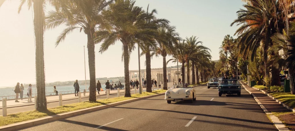Vintage cars driving along the palm-lined Promenade des Anglais on the French Riviera, under the afternoon sun.