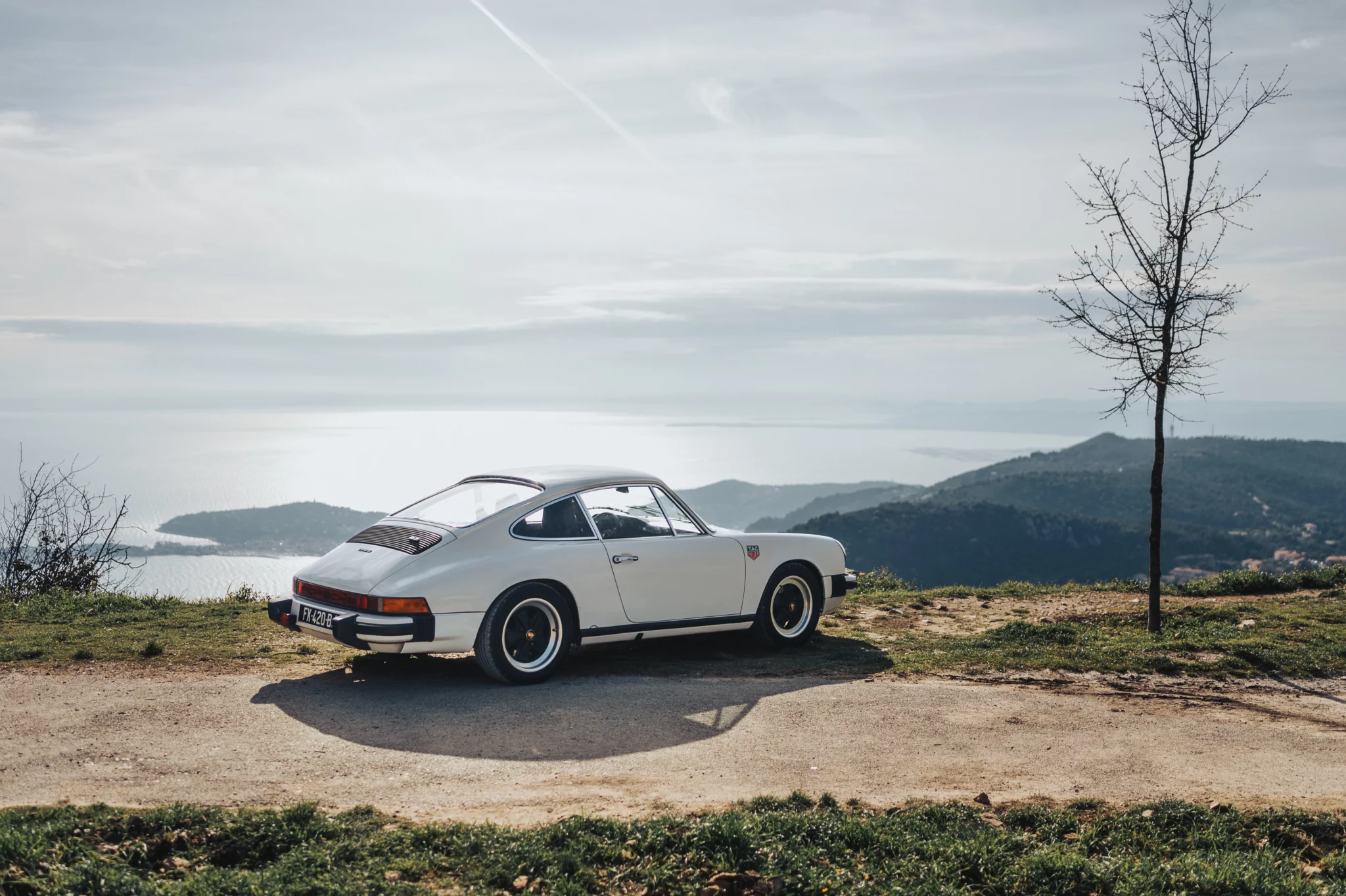 Vintage white Porsche 911 parked on a scenic viewpoint above the French Riviera coastline, with sea and mountains in the background.