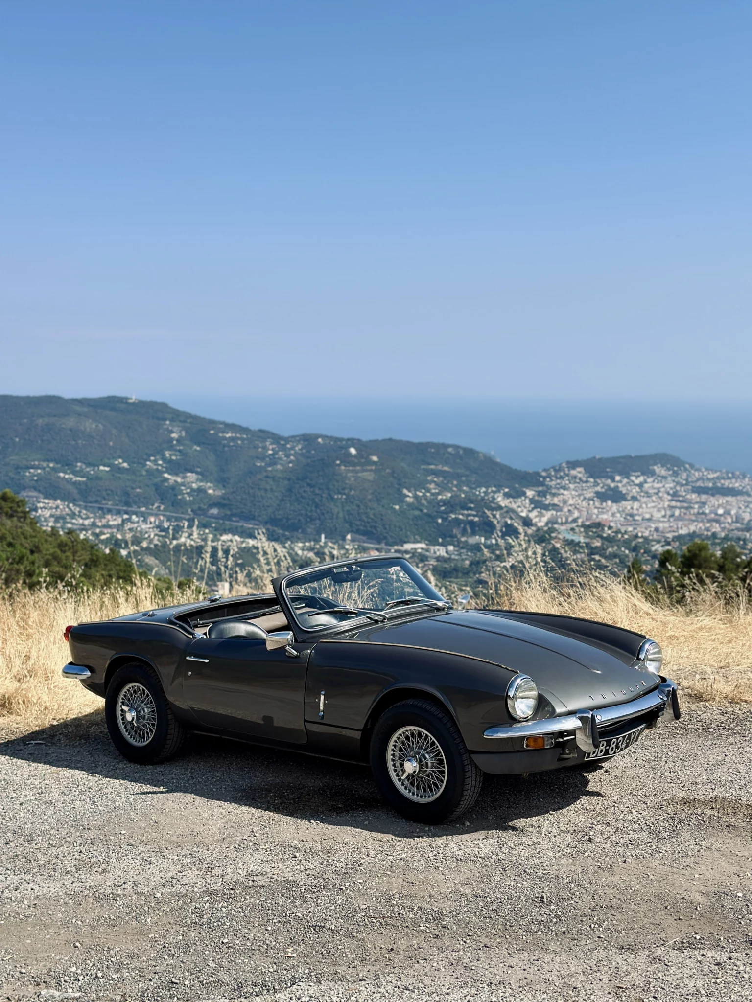 Triumph Spitfire convertible parked on a winding coastal road with the sea and distant hills in the background.