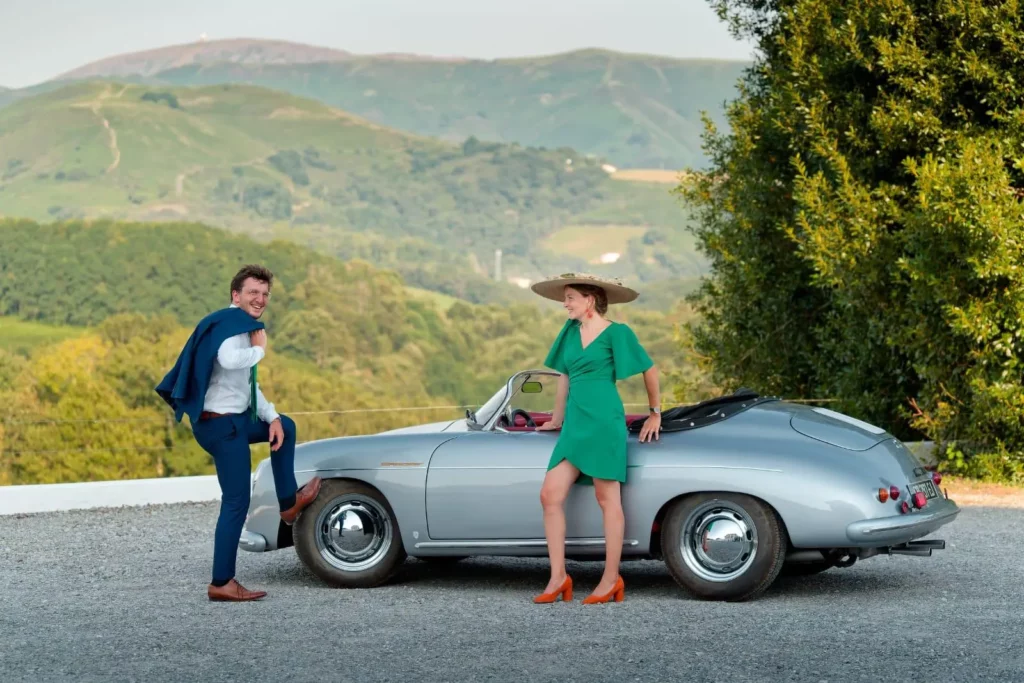 A stylish couple next to a silver vintage Porsche 356 convertible, set against the backdrop of the rolling green hills and La Rhune mountain in the Basque Country.