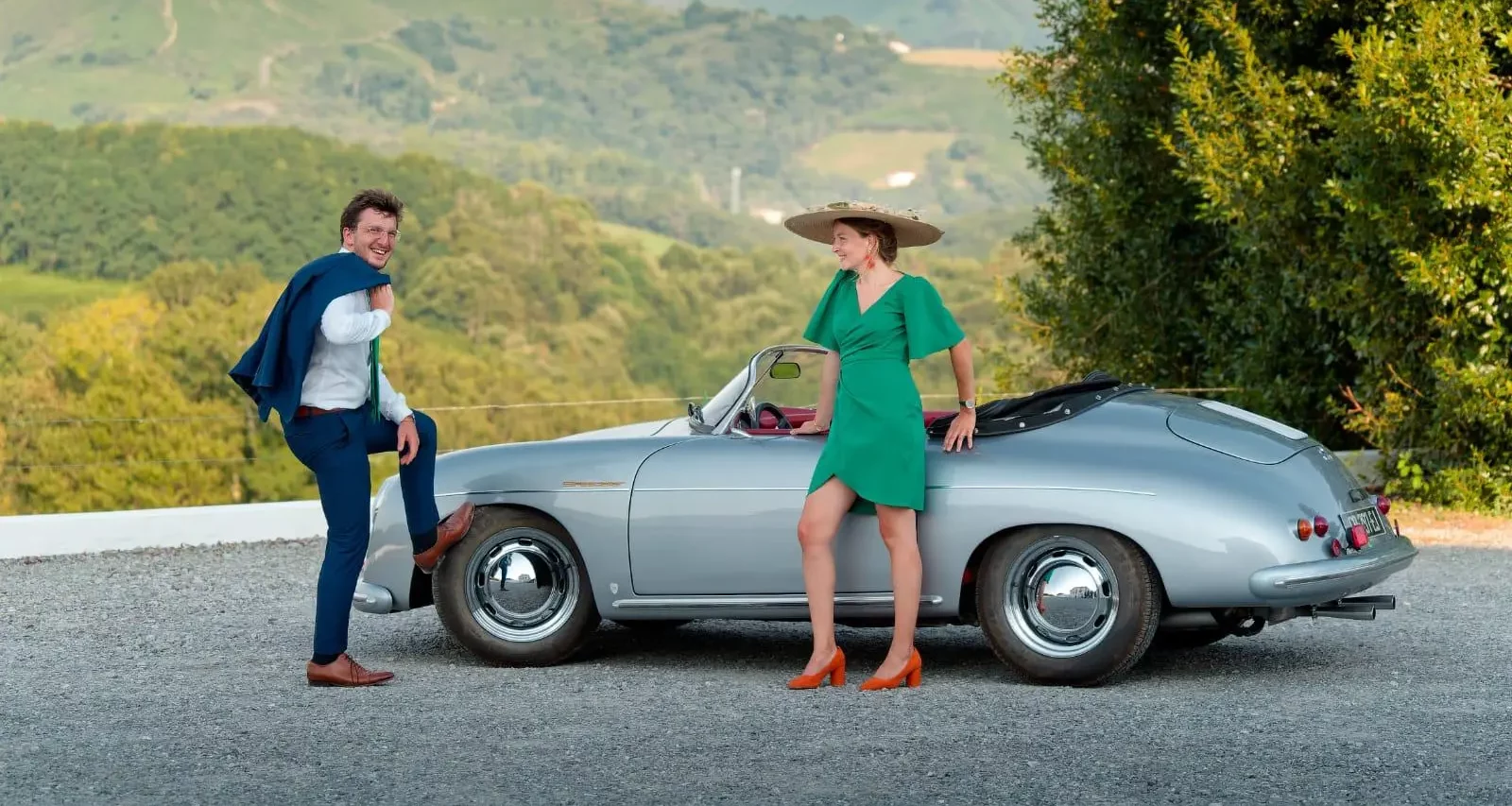 A stylish couple next to a silver vintage Porsche 356 convertible, set against the backdrop of the rolling green hills and La Rhune mountain in the Basque Country.