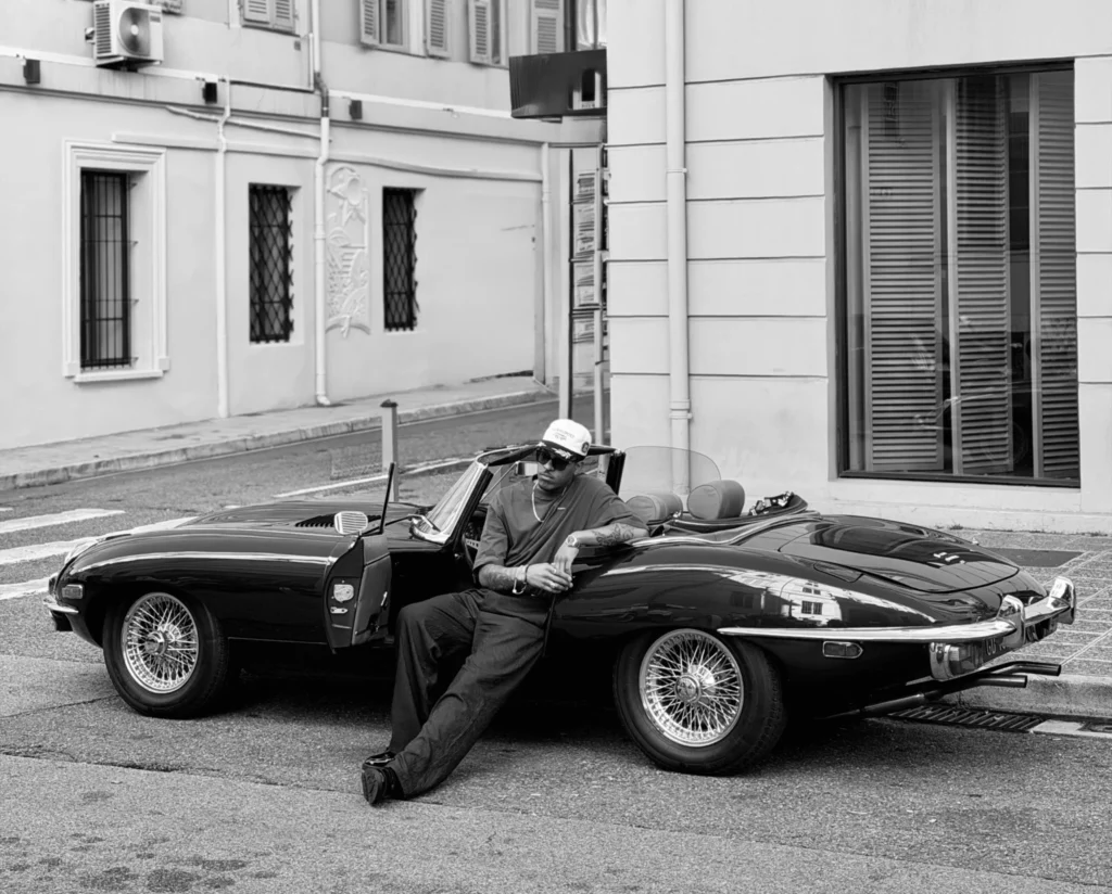 A man leaning against a vintage black Jaguar E-Type convertible on a street in Nice, France, wearing a stylish outfit and sunglasses, evoking a classic 007 James Bond aesthetic in front of Hotel Beau Rivage.