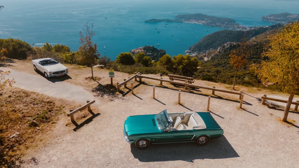 An aerial, high-angle view of two classic convertibles parked at a viewpoint on a gravel area overlooking the deep blue Mediterranean Sea on the Côte d'Azur. A vintage white Mercedes 280SL Pagode (top-left) and a polished turquoise 1965 Ford Mustang (center-right) are bathed in warm sunlight, surrounded by lush green and golden-autumn trees.
